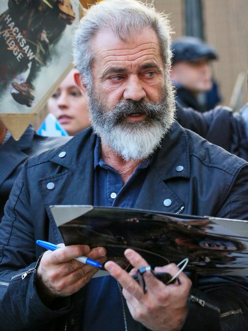 A close-up of a bearded older man in a black jacket signing an autograph with a blue pen, with a 'Hacksaw Ridge' poster visible in the background.