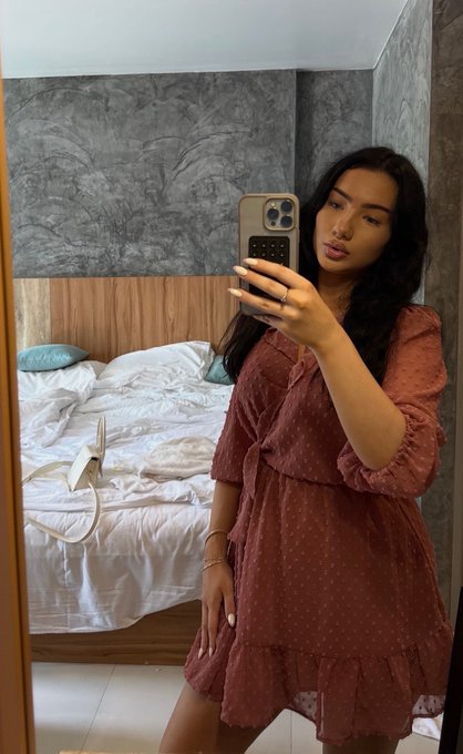 A young woman takes a mirror selfie in a bedroom, wearing a dusty rose swiss dot dress with a tiered hem and three-quarter sleeves. She holds up her phone while posing, with a messy white bed visible in the background, perfectly capturing a casual "GM" (good morning) vibe.
