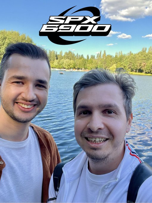 The image features two men standing next to each other, both smiling for a photo. They are positioned near a body of water, possibly a lake or river, with trees visible in the background. The caption of the image reads "First Moscow SPX meetup @vasiliy_spx6900 is very real & very legend as we all know Time to build SPX irl nodes globally $SPX #SPX6900". This suggests that the two men are attending a special event or gathering related to the SPX organization.