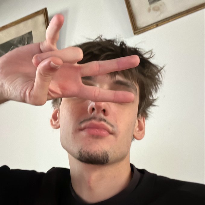 A young man with messy brown hair, a light mustache, and a goatee holds up two fingers in a casual pose, covering part of his face as he looks toward the camera in a low-angle selfie-style shot, with framed artwork visible on the wall behind him, fitting the relaxed "Yo GANG " vibe of the post.