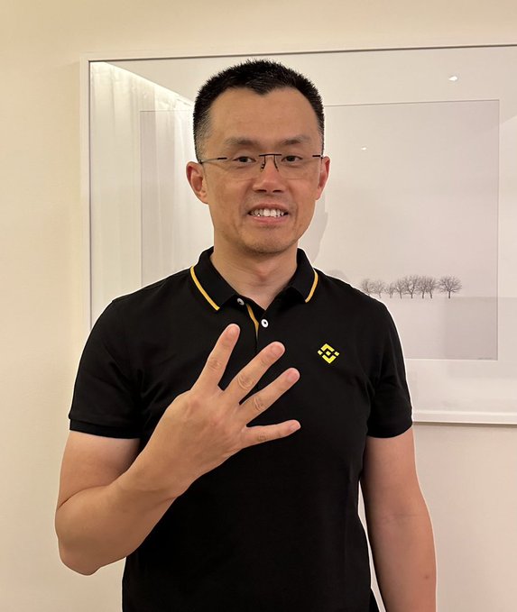 A man in a black polo shirt with a yellow Binance logo stands in front of a framed picture, holding up four fingers while smiling. There is no visible connection to Jayden Daniels in the image, but the Binance branding suggests he may be associated with the cryptocurrency exchange.