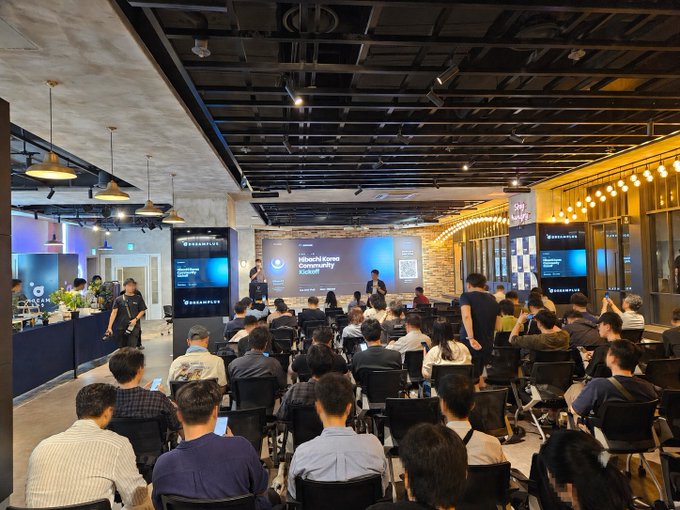 A wide shot of a conference or event titled 'Hibachi Korea Community Kickoff' taking place in a large room, with an audience seated in front of a stage and a presenter speaking.