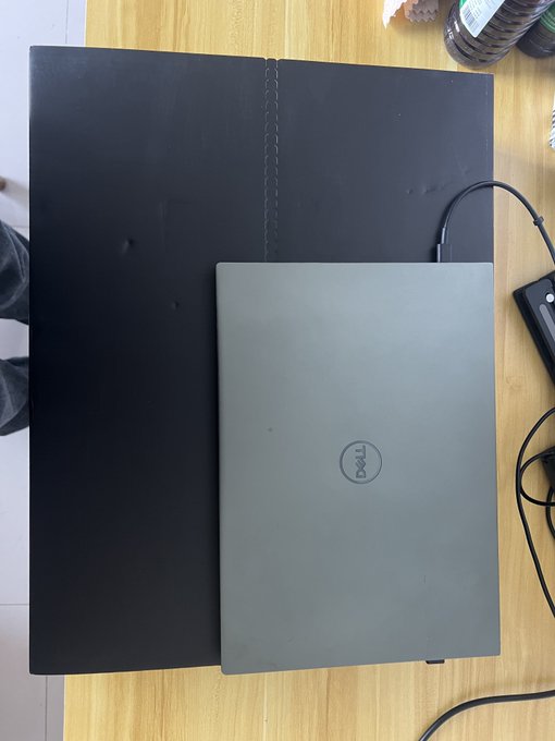 A closed silver Dell laptop sitting on top of a larger closed black laptop on a wooden surface, with cables and bottles visible nearby.