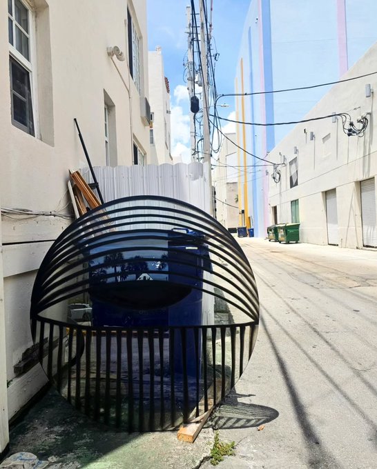 A black metal spherical art installation sits in an alleyway between white buildings, with a colorful geometric mural visible in the background.