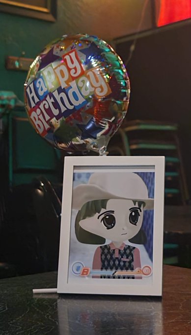 A "Happy Birthday" mylar balloon sits behind a framed photograph of the Vocaloid character Hatsune Miku's "Sharing the World" version, displayed on a table—fitting the post's birthday tribute to "miladys" (likely referring to Anime and Vocaloid fans).

Visible text includes "Happy Birthday" on the balloon and "Sharing the World" with "39" at the bottom of the framed photo.
