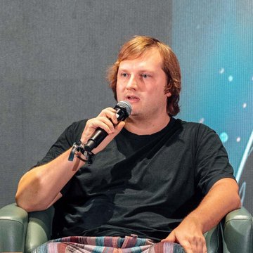 A young man with reddish-blonde hair, wearing a black t-shirt and beaded bracelets, speaks into a microphone while seated in a teal armchair at what appears to be a panel discussion.