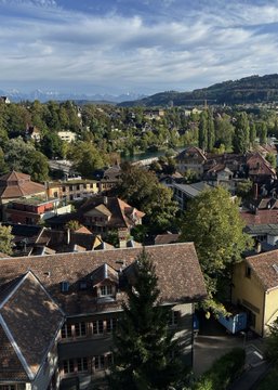 An aerial view of a town with red-tiled roofs, green trees, and distant mountains under a partly cloudy sky.