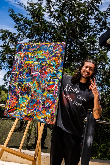 A smiling man with long dark hair poses with a peace sign next to an easel displaying a vibrant, abstract, multicolored painting; he wears a black t-shirt that reads 'Kinda Cute Kinda Don't'.