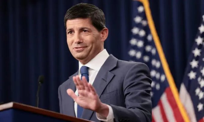 A man in a dark suit and blue tie gestures with his hand while speaking at a podium, with American flags displayed in the background.