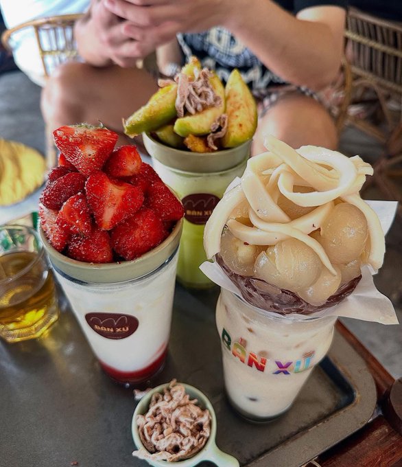 Three elaborate desserts from 'ĂN XUẤT' topped with fresh strawberries, figs, and jackfruit, served with a small dish of chocolate.