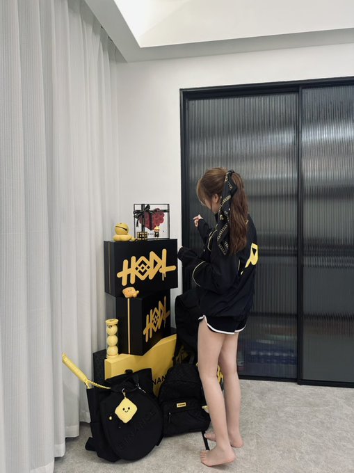 A young woman stands barefoot in her bedroom arranging a dedicated Binance display corner, organizing black and yellow boxes marked with "HEYI" branding alongside plush toys, a framed item, and Binance-branded bags on the floor.

The image illustrates the post's caption about setting up a Binance-themed corner, showing the early assembly stage with branded merchandise and decorative items being arranged into what will become a striking home display.