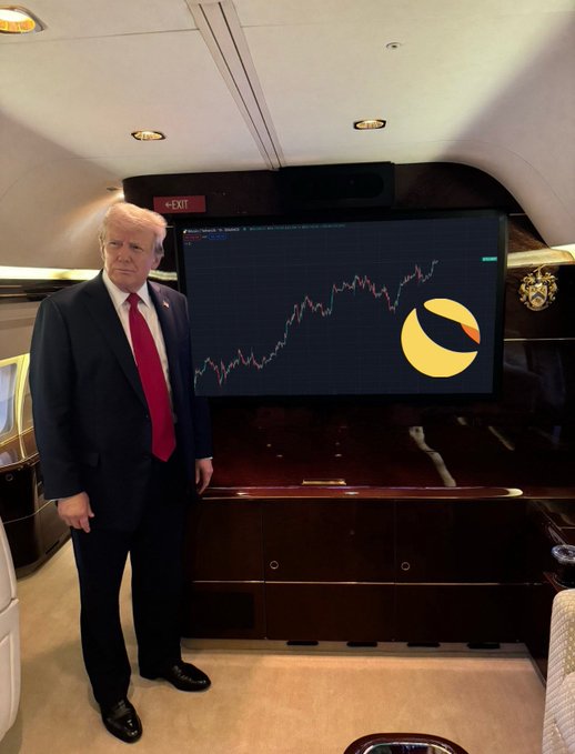 A man dressed in a suit, wearing red tie, stands in front of a large screen displaying a stock chart with the Trump's name on it and the words "buy $LUNC" above him.