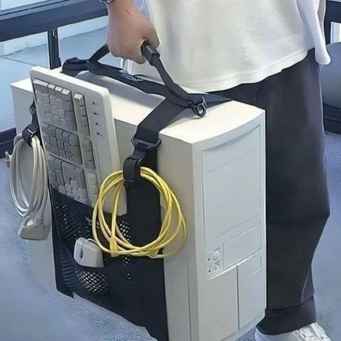 A person carries a large beige desktop PC tower by a strapped handle, with a keyboard tucked under the top strap and a coiled yellow cable and a white cable hanging from the side—matching the post's description of lugging a full-sized desktop computer on the go for AutoCAD drafting work.