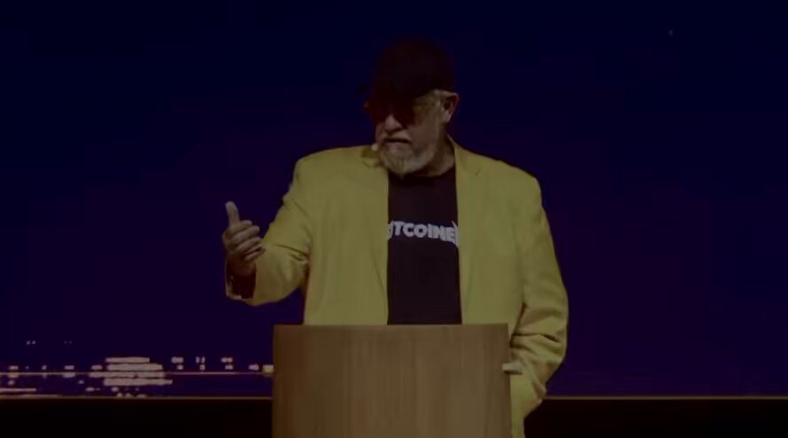 The image shows a man standing at a podium, giving a speech or presentation. He is wearing a yellow jacket and sunglasses, and he has his hand raised above his head as if emphasizing a point during his talk. The caption of the image reads "Go watch the full video of the BTC Prague talk from @DrJackKruse now". This suggests that there might be an event related to Bitcoin or cryptocurrency taking place in Prague, possibly at a conference or gathering.
