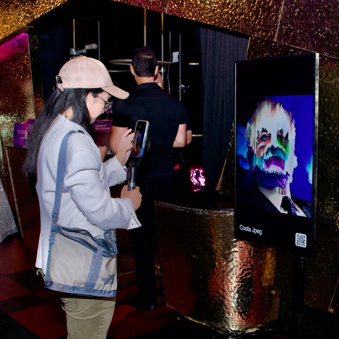 A woman wearing a cap and light blue blazer holds up a smartphone at what appears to be an art exhibit, standing near a digital display featuring a glitchy portrait labeled "Costa Jpeg" with a QR code.