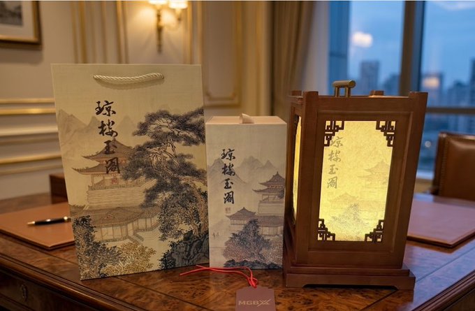 The image features a wooden desk with three items displayed on top of it, including two lanterns and a paper bag. The lanterns are positioned at different angles, creating an interesting visual effect. A note attached to one of the lanterns reads "市场暴跌 仓位被套 本以为币场中感受到了一丝久这个灯笼摆着真的挺好看 提前祝大家端午安康 。 @Heidi_MGBX". The post seems to be related to the lanterns, possibly discussing their design or origin.