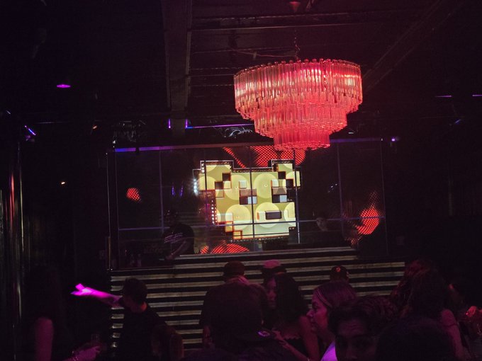 A photograph of a dark nightclub interior featuring a large illuminated red chandelier hanging above a digital screen, with patrons gathered below.