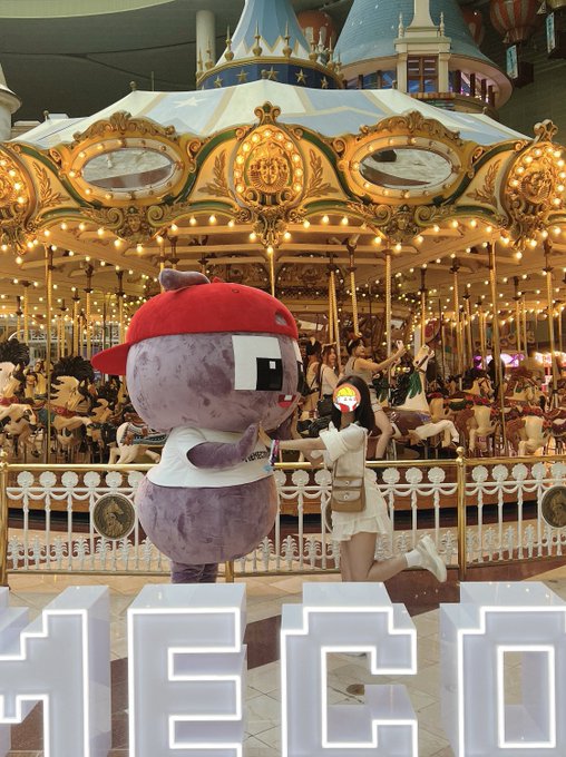 A purple mascot character with a red cap and an eye patch poses with a woman in a white dress in front of an ornate, illuminated carousel inside what appears to be a shopping mall.