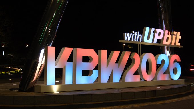 A nighttime photo shows a large illuminated sign reading 'KBW2026 with Upbit' in red, white, and blue colors.