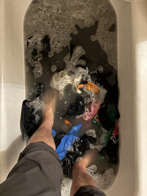A first-person view of someone soaking their bare feet in a bathtub filled with murky, soapy water and various articles of clothing.