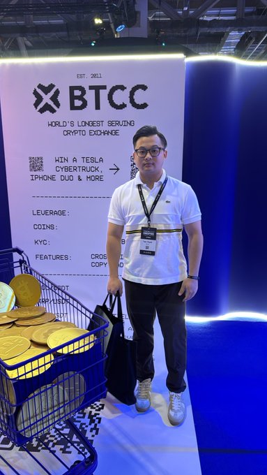 A man wearing a white polo shirt, glasses, and an event badge stands at a BTCC booth beside a cart of gold-colored physical crypto coins. The backdrop reads “EST. 2011,” “BTCC,” “WORLD’S LONGEST SERVING CRYPTO EXCHANGE,” “WIN A TESLA CYBERTRUCK, IPHONE DUO & MORE,” and lists features including “LEVERAGE,” “COINS,” and “KYC.”
