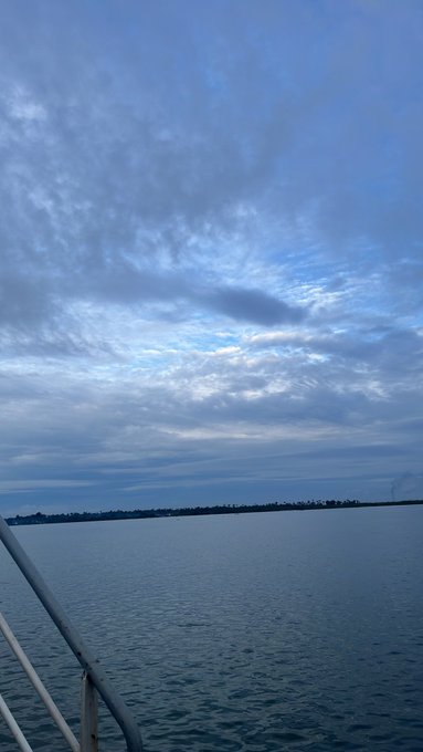 A serene view of a body of water under a cloudy blue sky, taken from a boat with metal railings visible in the foreground.