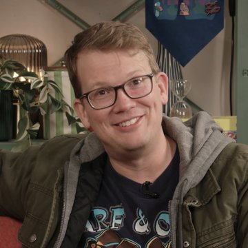 A portrait of a smiling man with reddish-brown hair wearing glasses and a green jacket over a graphic t-shirt.