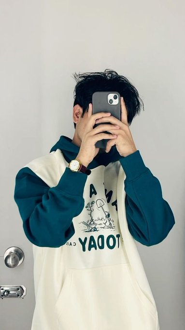 A mirror selfie of a person wearing a cream and teal hoodie with 'TODAY' printed on it, holding a phone that covers their face.