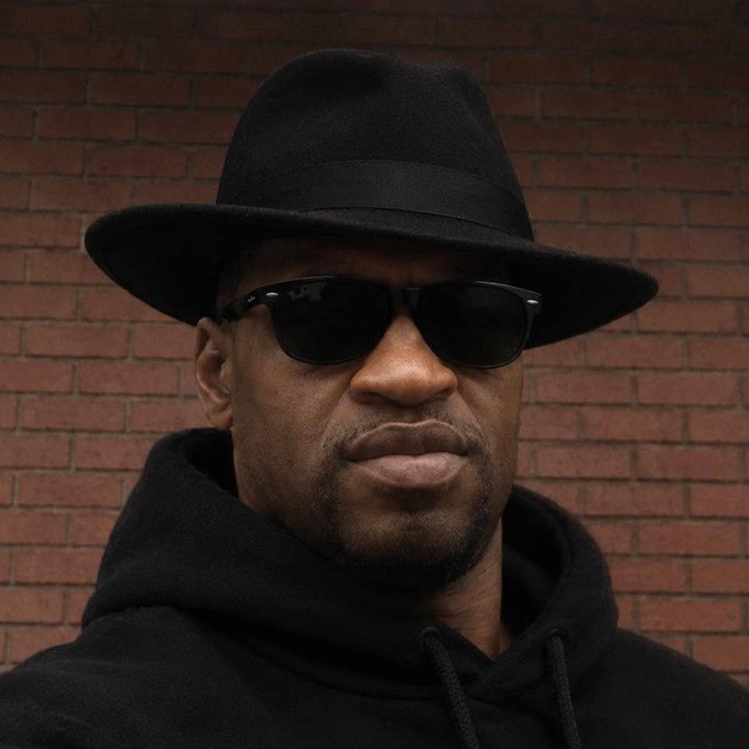 A stylish man wearing a black fedora, dark sunglasses, and a black hoodie poses against a red brick wall, exuding a confident "Gm" (good morning) vibe to start the day.