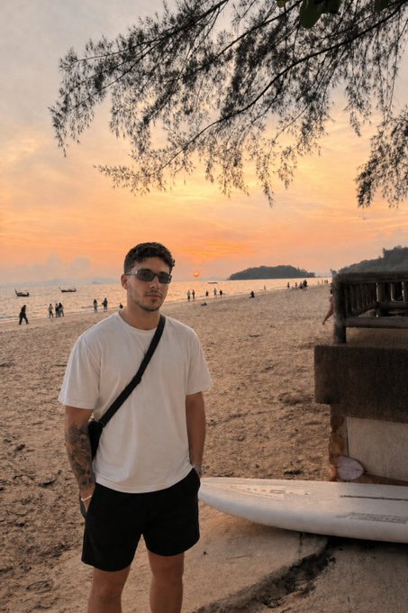 A young man wearing sunglasses and a white t-shirt stands on a beach at sunset, posing next to a surfboard with golden skies and distant islands in the background. The beachfront setting aligns perfectly with the post's casual, oceanic vibe, complementing the request for a good morning greeting and a crypto wallet address.
