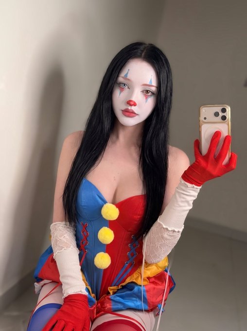 A woman taking a mirror selfie in a jester/clown costume featuring a red and blue corset with yellow pom-poms, white face makeup with red accents, and long red satin gloves, holding an iPhone with a gold case.