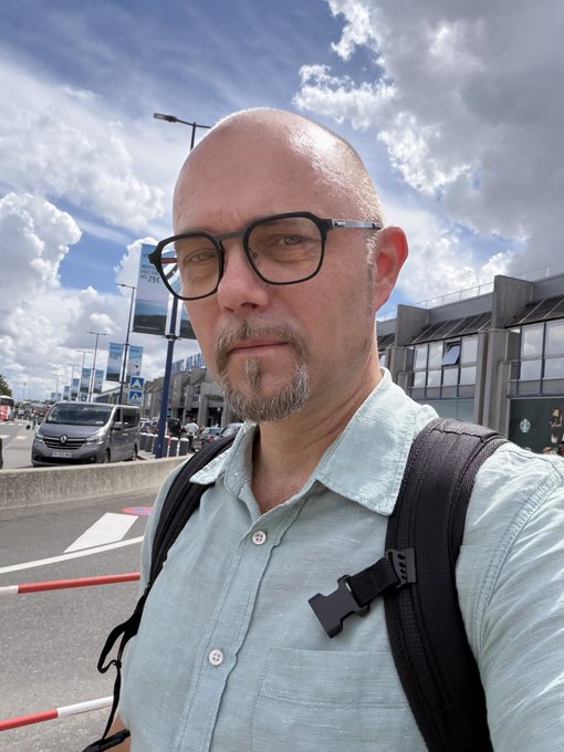 Selfie of a bald man with glasses and a goatee wearing a light green shirt and backpack straps, taken outdoors near a street with cars.