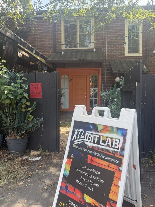 An exterior photo of "ATL BitLab" - Atlanta's Freedom-Tech Hackerspace - showing a sandwich board listing services like tech meetups, co-working office, event space, and maker tools, placed in front of a black gate and brick building.