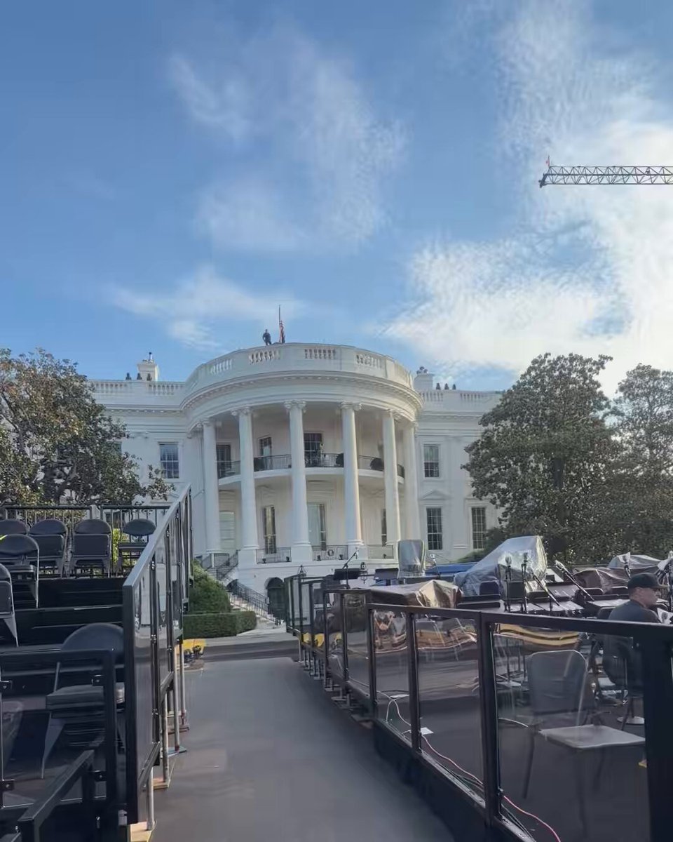 The image shows a white building with columns, which appears to be the White House. The photo has been edited to include a black fence surrounding the building, giving it an artistic touch. In addition to the main subject, there are several chairs placed around the area and a person standing near one of them. A construction crane is also visible in the background, suggesting that some work or development might be taking place nearby. The post accompanying this image provides information about the location and context of the scene.