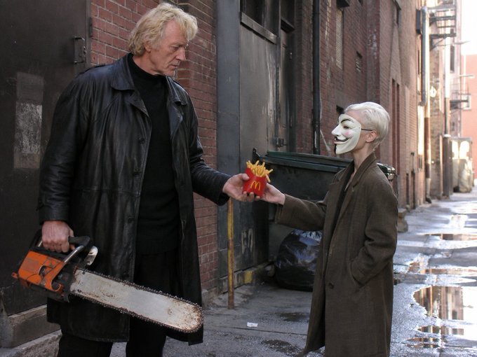 A man in a black leather coat holds a chainsaw and hands a McDonald's french fries box to a person wearing a Guy Fawkes mask and brown coat in an alleyway.
