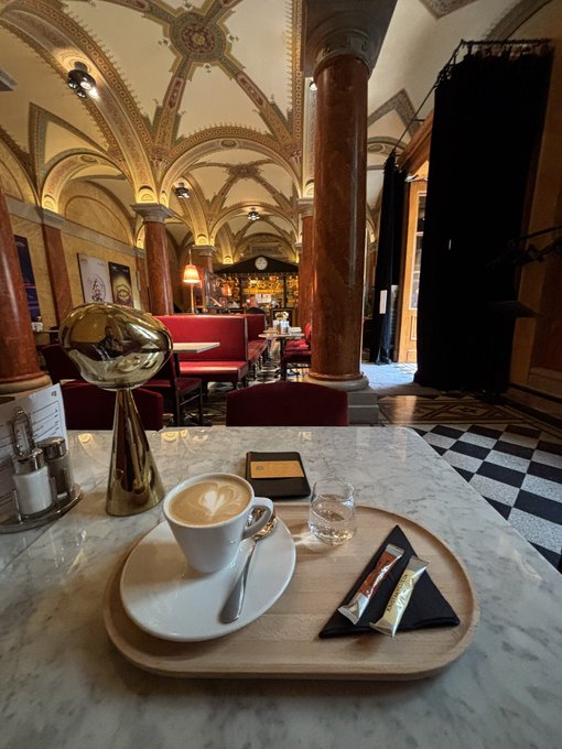 A cappuccino with latte art and two wrapped candies sit on a wooden tray atop a marble table inside an ornate, vaulted-ceiling cafe with classical columns and red velvet seating.