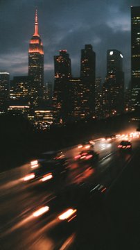 A nighttime cityscape shows illuminated skyscrapers and streaks of car lights from traffic on a busy street below.