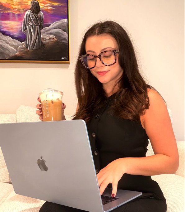 A woman wearing glasses sits with an open Apple laptop while holding an iced drink; a framed painting hangs behind her on the wall.