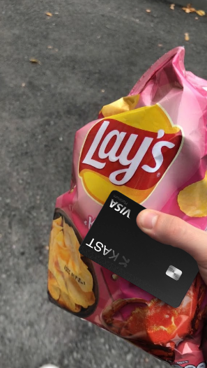 A hand holding a pink bag of Lay's potato chips along with a black credit card while standing on asphalt pavement.