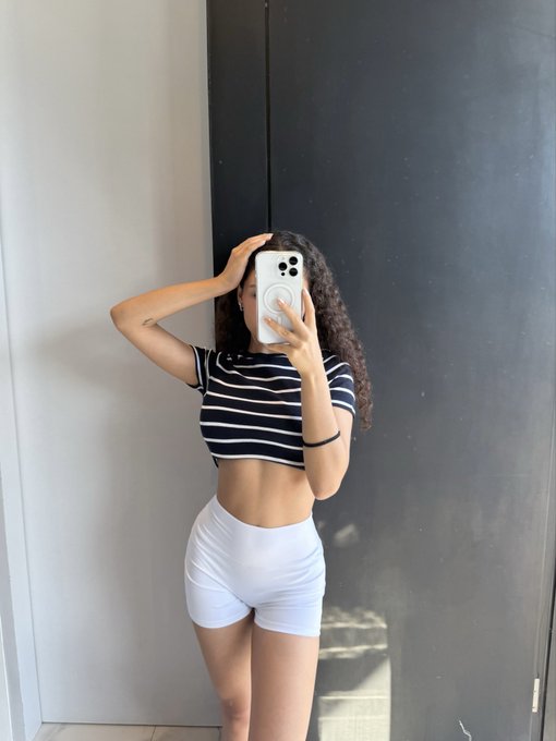 A mirror selfie of a young woman with curly hair wearing a black and white striped crop top and white shorts, holding a smartphone in front of her face.