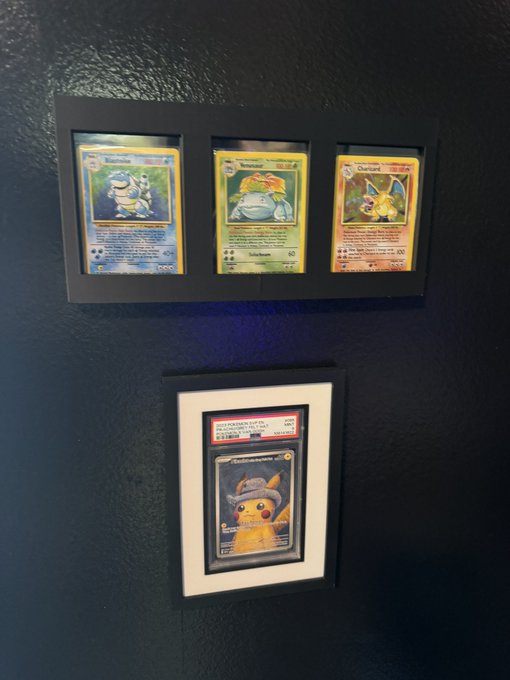 Four framed Pokémon trading cards displayed on a dark blue wall, including what appears to be Blastoise and Charizard cards in a horizontal triple frame above a single PSA-graded Raichu card.