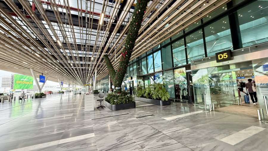 An interior view of a modern, spacious airport terminal with high ceilings, large windows, decorative plants, and gate signs including 'D5' visible.