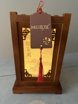 The image features a wooden lantern with a red tassel hanging from its handle, giving it an elegant appearance. The lantern has a tag attached to it that reads "MGBX", which could be a brand or logo associated with the lantern's design or function. The lantern is placed on a table and appears to be made of wood, possibly crafted by a skilled artisan.