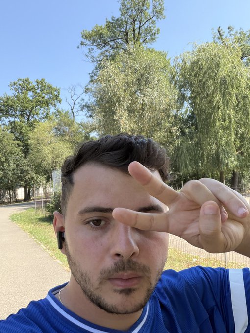 A young man with dark hair and an earpiece takes a selfie outdoors in a park, holding his fingers in a peace sign over his eye, with trees and a paved walkway visible behind him.