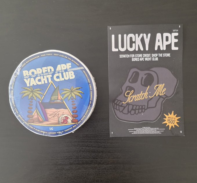 Two 'Bored Ape Yacht Club' branded promotional items on a dark surface: a round tin labeled 'Bored Ape Yacht Club' and a scratch-off card titled 'LUCKY APE' with a 'Win up to $500' starburst.