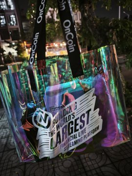A photograph of an OnChain event tote bag with holographic, iridescent material containing promotional materials, with the tagline 'THE WORLD'S LARGEST LIVE STREAMING PLATFORM' visible on the contents.