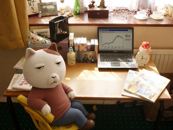 Quirky scene of a plush cat toy character wearing a red sweater and blue jeans, sitting at a wooden desk with a Samsung laptop displaying a stock chart showing '$ket' with an upward trend, plus various desk items.