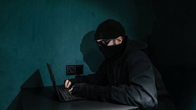 A masked person in a black beanie, hoodie, and gloves types on a laptop at a desk against a teal wall, lit by a narrow beam of light.