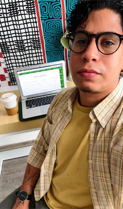 A young man with glasses and curly hair wearing a yellow t-shirt and plaid overshirt takes a selfie in a cafe, with a laptop showing 'applestheobie' on the screen and a coffee cup on the table beside him.