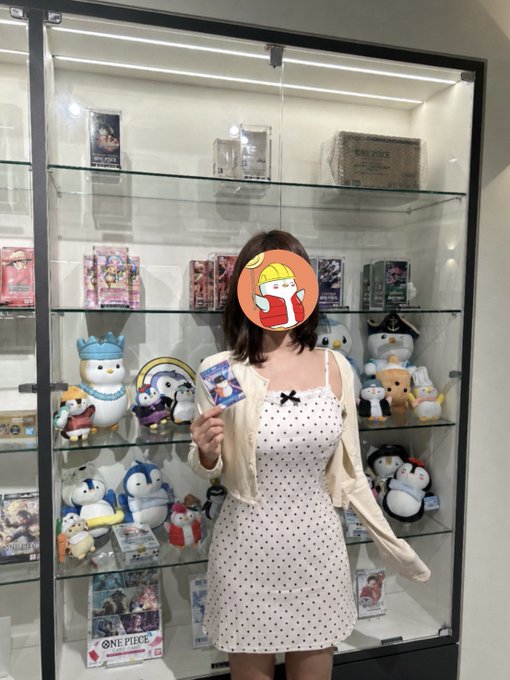 A young woman in a white polka-dot dress with a cream cardigan standing in front of a glass display case filled with One Piece merchandise and plush toys, holding a small card with a cartoon sticker covering her face.